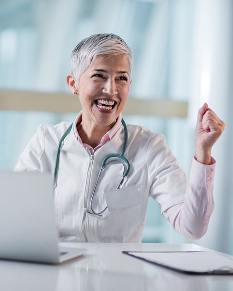 Excited medical resident, white woman with short silver hair, looks up from laptop and pumps fist. 