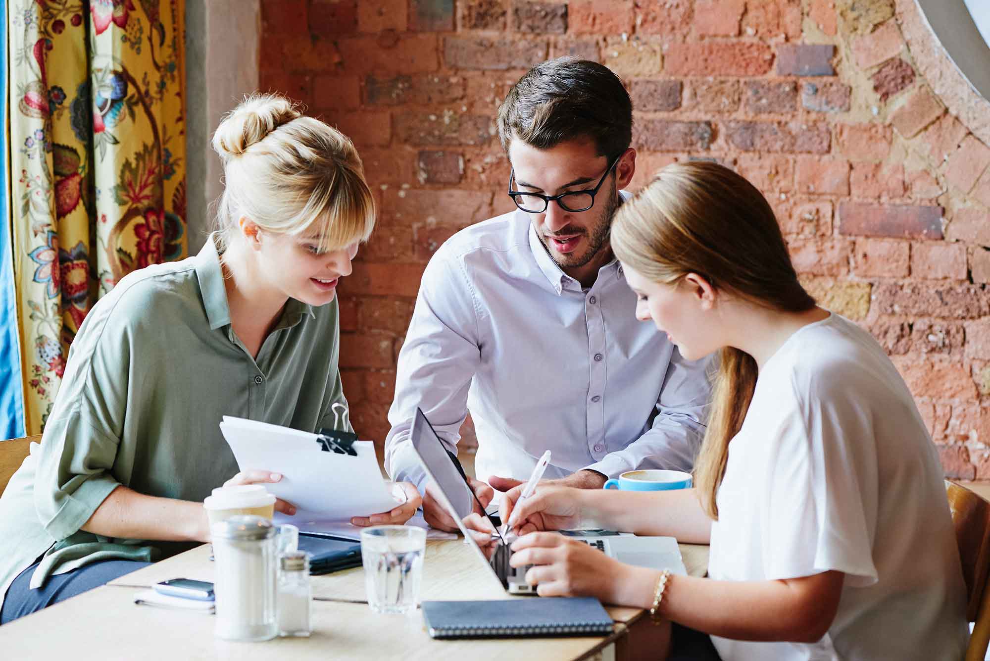 Two white women with blonde hair and a white man with brown hair, beard and wearing glasses, at a high-top table making plans. 